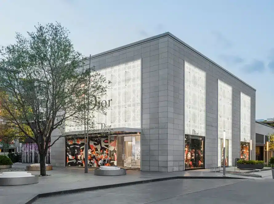A modern Dior store with a light gray exterior, large illuminated display windows, and minimalist architectural design, located in an open plaza with trees and benches nearby.