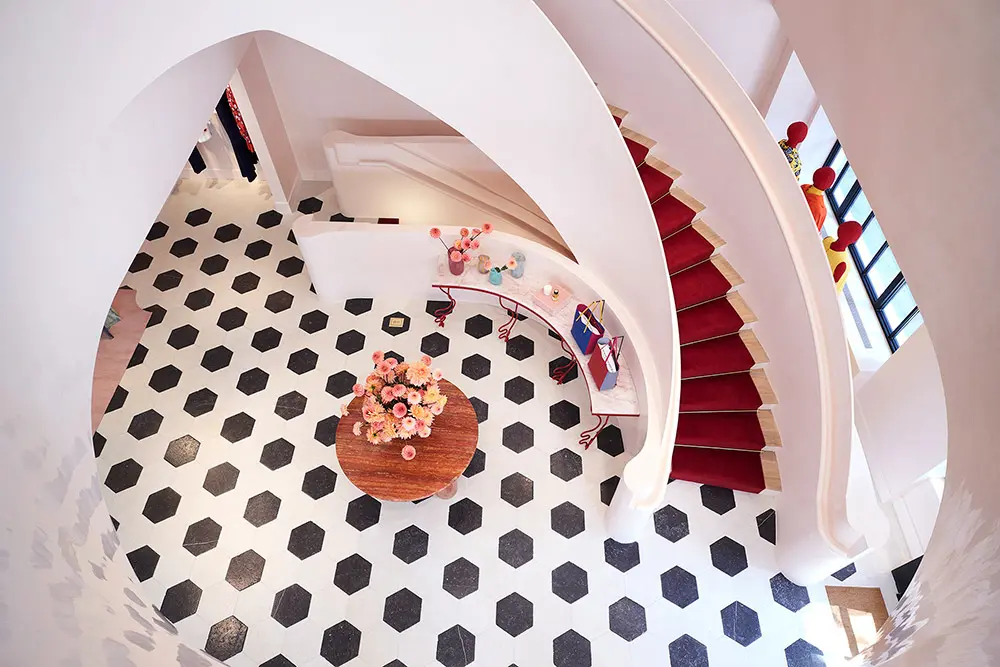 A spiral staircase with red carpet curves around a round table topped with a flower arrangement. The floor features a black and white hexagonal tile pattern and the space is bright with natural light.