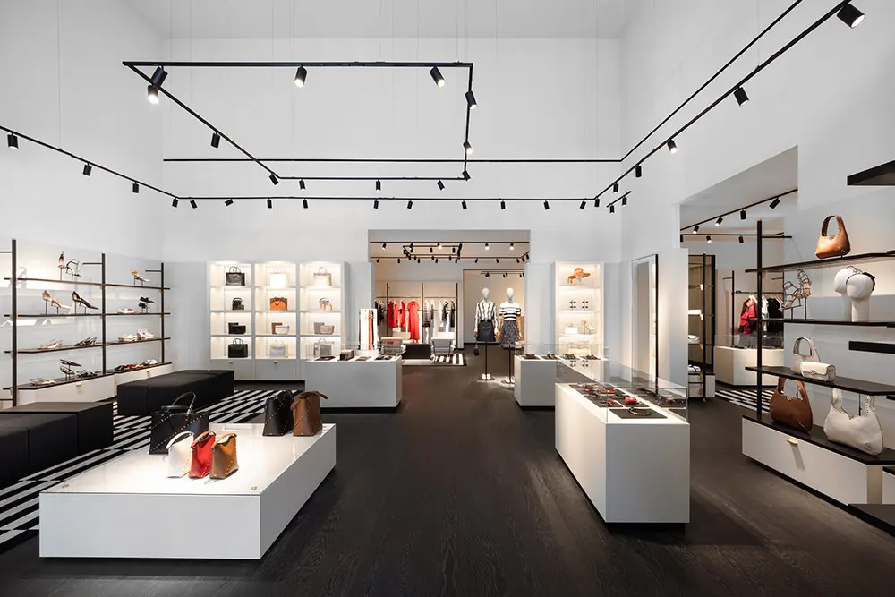 Modern fashion boutique with white walls, black flooring, and minimalist lighting. Display shelves and tables showcase handbags, accessories, and clothing. The space is open, well-lit, and organized with a sleek, stylish design.