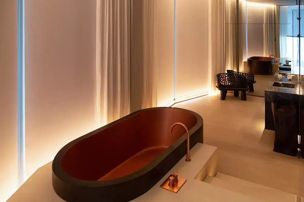 A modern bathroom with soft lighting, featuring a brown freestanding bathtub with a copper faucet, light-colored floors, curtains, a black woven chair, and a large wall mirror reflecting the space.