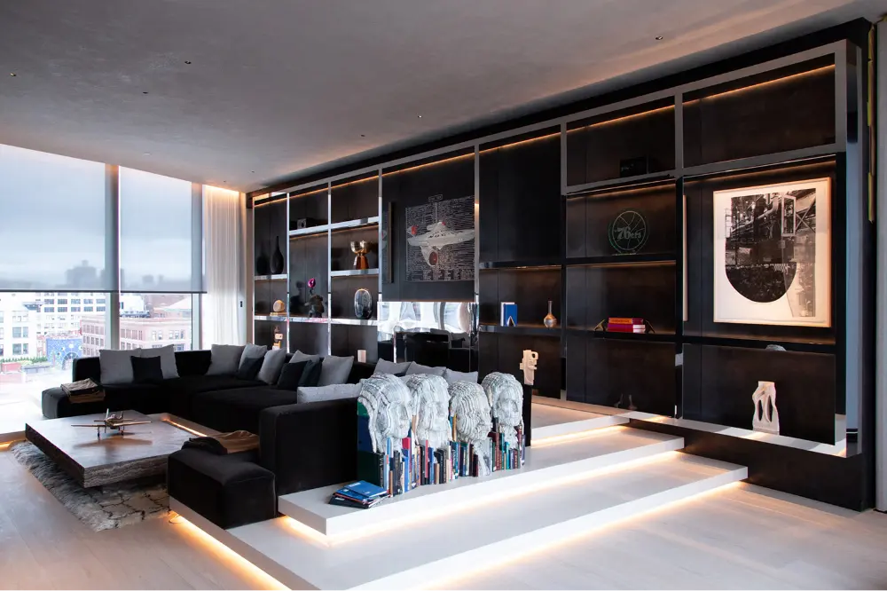 Modern living room with a large black sectional sofa, built-in bookshelves, ambient lighting, and floor-to-ceiling windows offering a city view. Artworks and books decorate the shelves and a blanket covers the sofa.