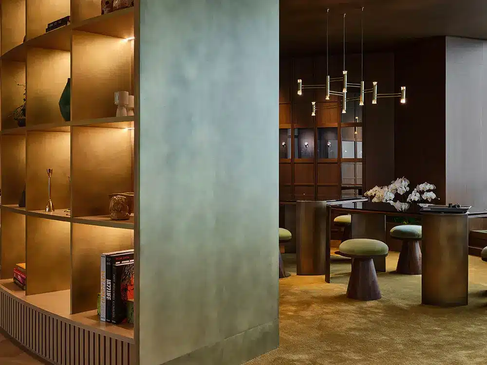 A modern interior with a large, illuminated shelving unit displaying decor items and books. On the right, there’s a dining area with round stools, a table, orchids, and a contemporary chandelier overhead.