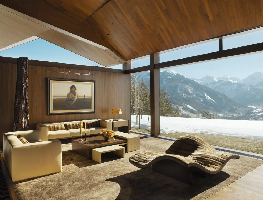 Modern living room with large floor-to-ceiling windows showcasing a snowy mountain view. Inside, there are cream-colored sofas, a curvy lounge chair, a coffee table, a lamp, and a painting on a wood-paneled wall.