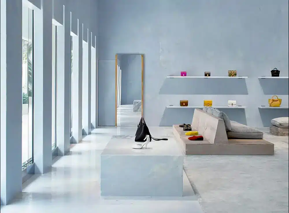 A modern, minimalist boutique with light blue walls, large windows, white floors, and open shelving displaying colorful handbags. A sofa sits in the center, and a black handbag is on a white display table.
