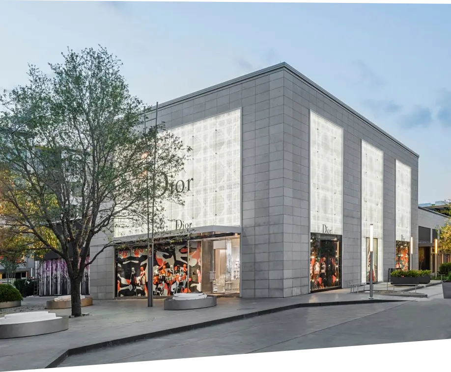 A modern, minimalist Dior store with large illuminated windows and a light gray facade sits on a clean, empty street. A tree and benches are visible in the foreground.
