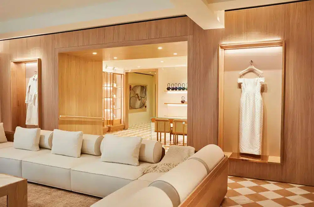 A modern, elegant lounge with light wood walls, a cream sectional sofa with white pillows, and display alcoves featuring a white dress on a hanger. The space is softly lit with a stylish, minimalistic decor.