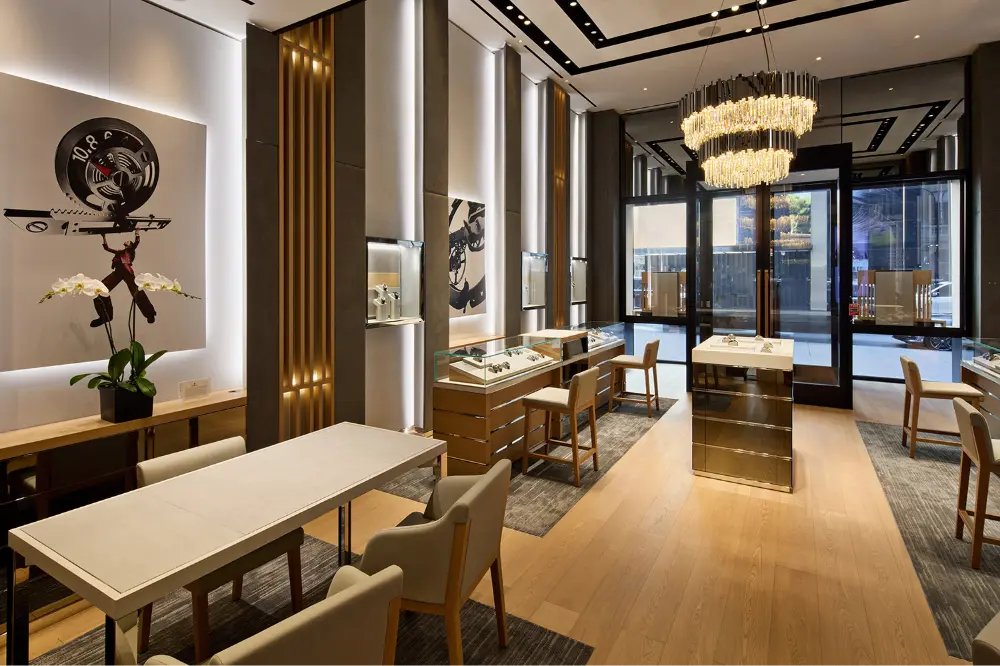 Modern, elegant jewelry store interior with sleek display cases, wooden floors, beige chairs, a large chandelier, wall art, and orchids. Natural light enters through glass doors and windows at the front.