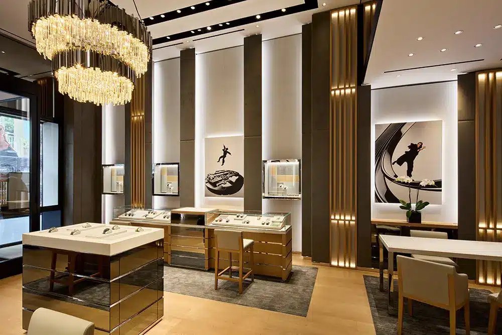 A modern, elegant jewelry store interior with gold accents, illuminated display cases, chandelier lighting, artwork on the walls, and several tables and chairs for customers.