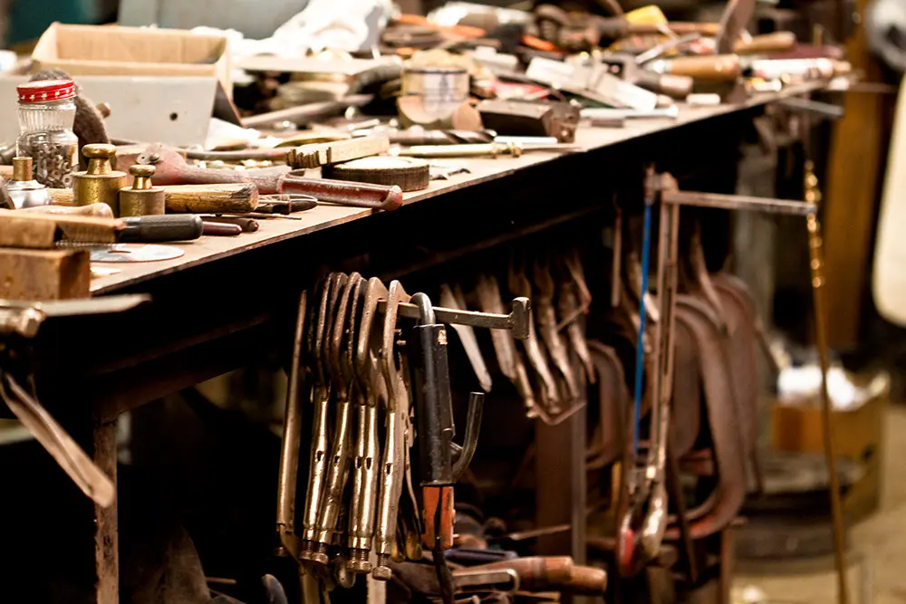 A cluttered workbench covered with various hand tools, jars, and parts, with several metal clamps hanging from the table's edge in a workshop setting.