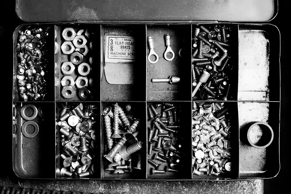 A black and white photo shows an open compartmentalized toolbox with various screws, washers, nuts, and small hardware pieces neatly sorted in each section.
