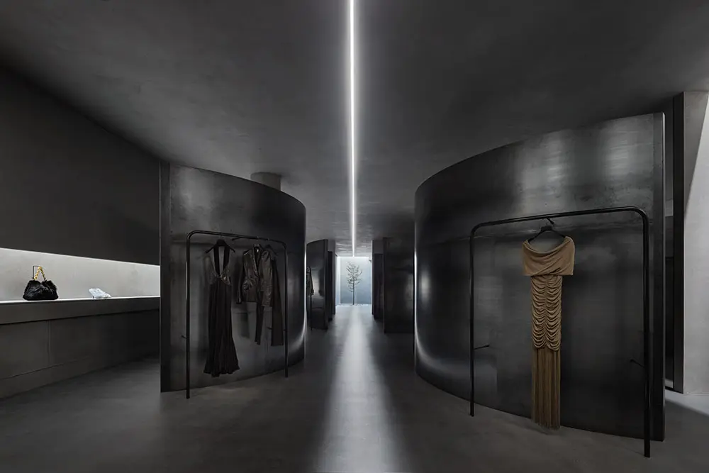 A modern, minimalist clothing store with curved black partitions, dim lighting, and a central LED strip on the ceiling. A beige dress and dark outfits are displayed on racks, with bags and accessories on a side shelf.