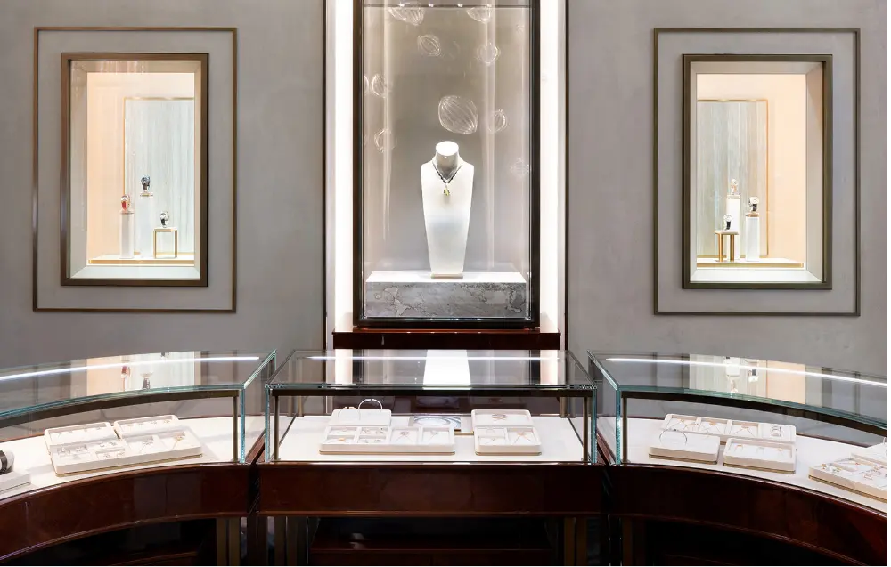 A luxurious jewelry store interior featuring glass display cases with jewelry and a central illuminated showcase displaying a necklace bust. Two framed niches with jewelry are mounted on the wall in the background.