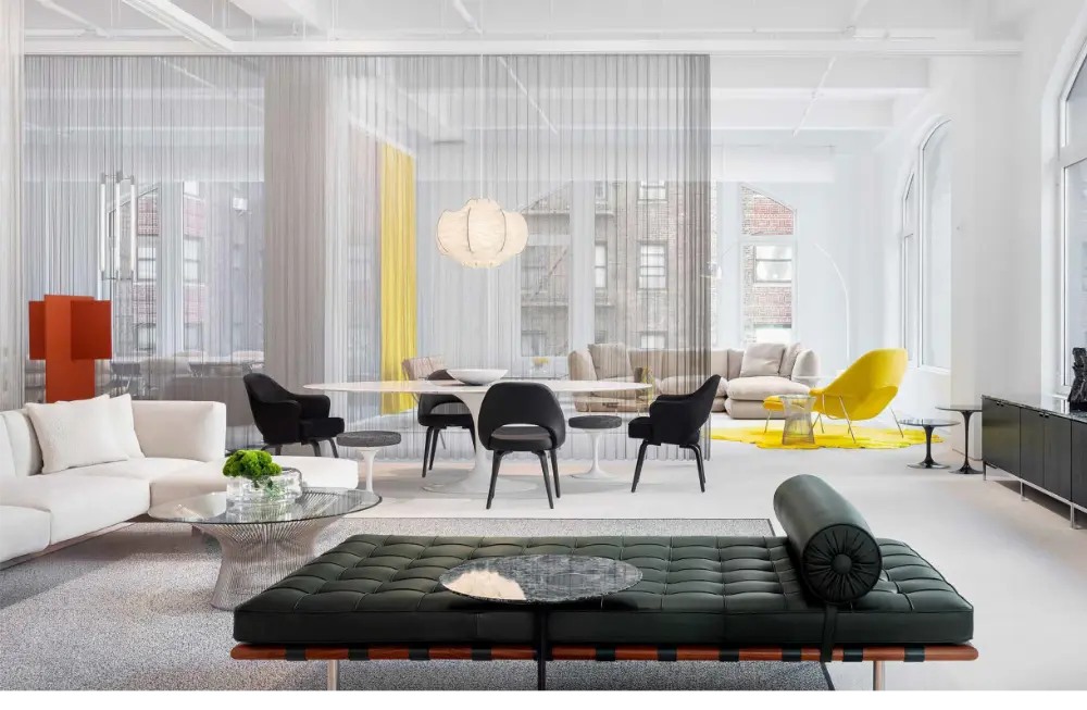 A modern, open-concept living space with white walls, large windows, sleek furniture, a black leather bench, white sofa, glass coffee tables, black and yellow chairs, and a sheer curtain dividing the room.