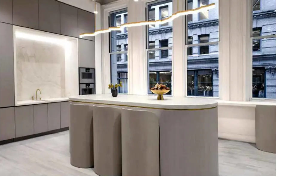 A modern kitchen with a large oval island, light gray cabinetry, built-in appliances, and tall windows overlooking a city street. A fruit bowl sits on the island under a wavy, minimalist ceiling light.