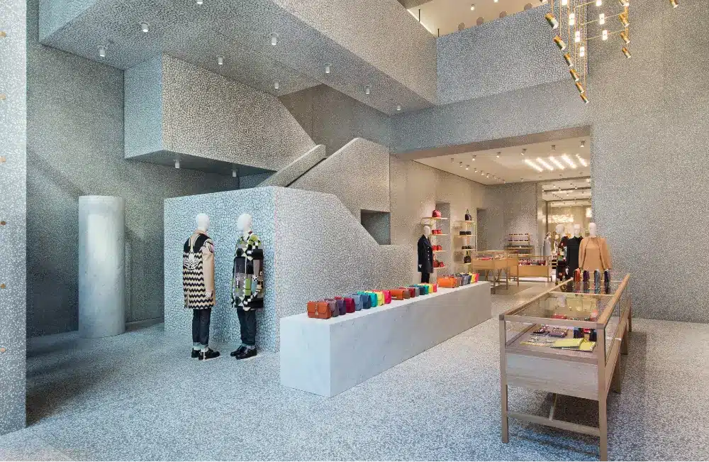 A modern, minimalist clothing store with mannequins dressed in designer outfits, shelves and tables displaying colorful accessories, and a geometric staircase in a spacious, light-filled interior.
