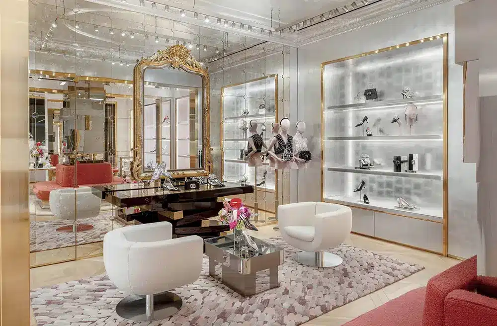 Luxurious boutique interior with gold-framed mirror, glass shelves displaying designer accessories, white modern chairs, a patterned rug, and fresh flowers on a sleek coffee table. The space has elegant lighting and reflective surfaces.