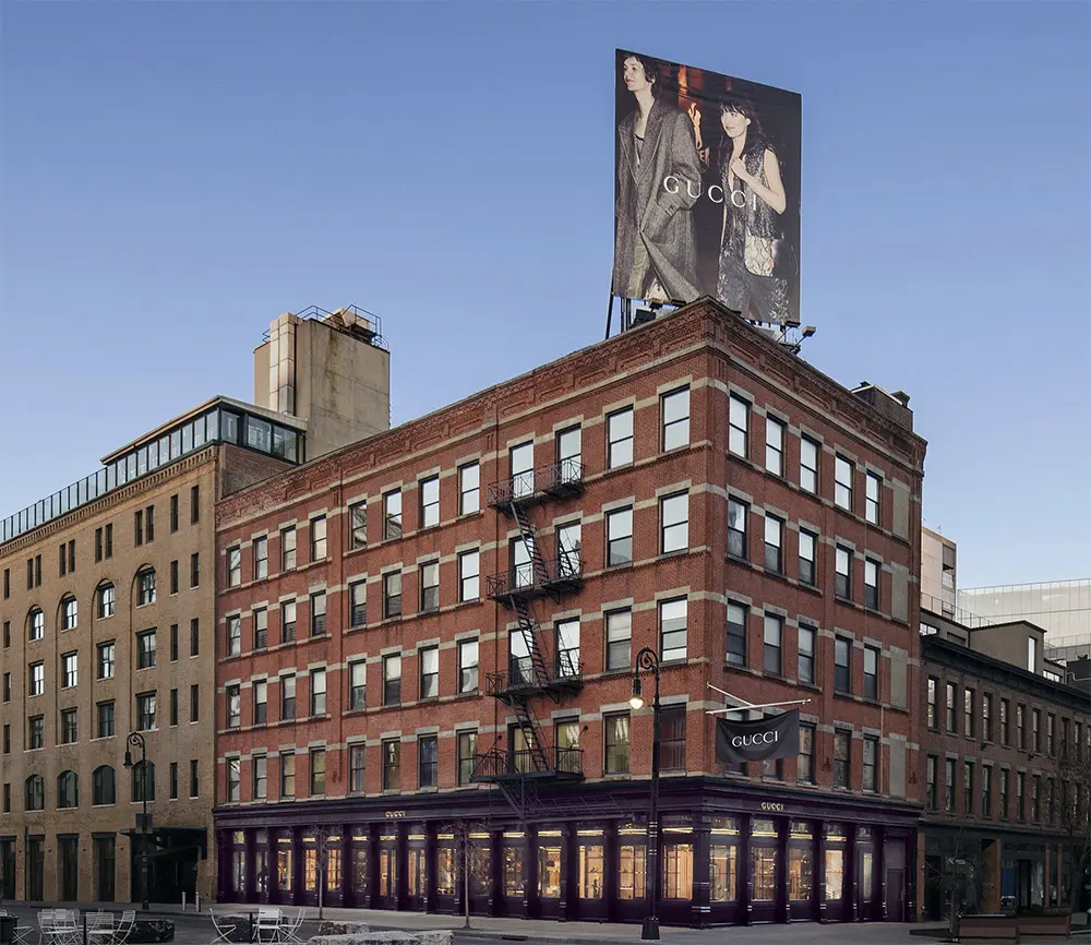 A four-story brick building with a Gucci store on the ground floor, outdoor seating nearby, and a large Gucci billboard featuring two models on the roof, against a clear blue sky.