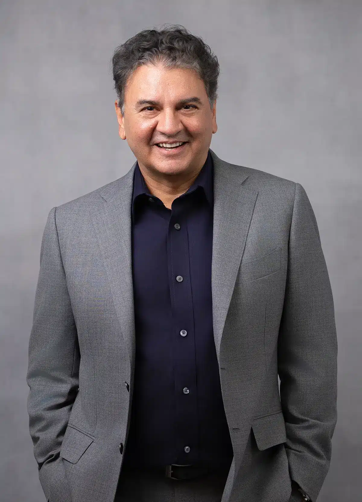 A man with dark, wavy hair wearing a gray suit jacket over a dark navy button-up shirt stands smiling against a plain gray background.