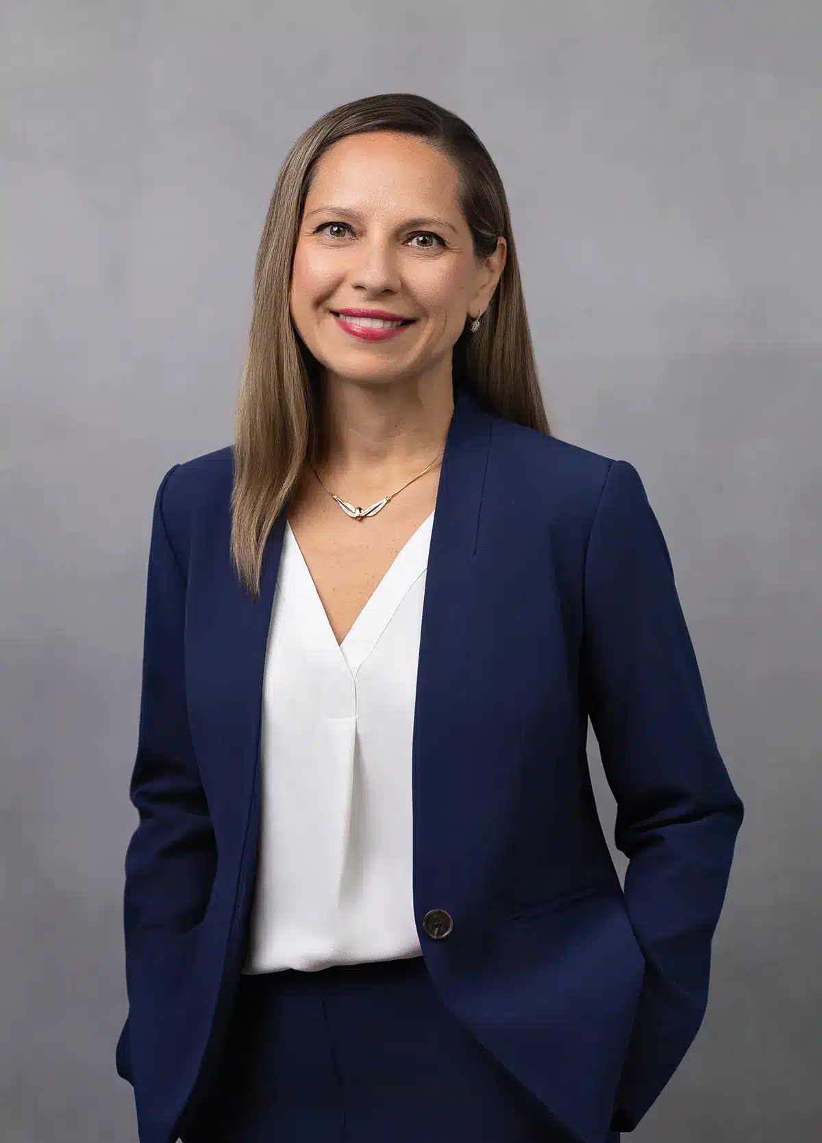 A woman with straight, light brown hair, wearing a navy blue blazer over a white blouse, smiles while standing against a plain gray background with her hands in her pockets.