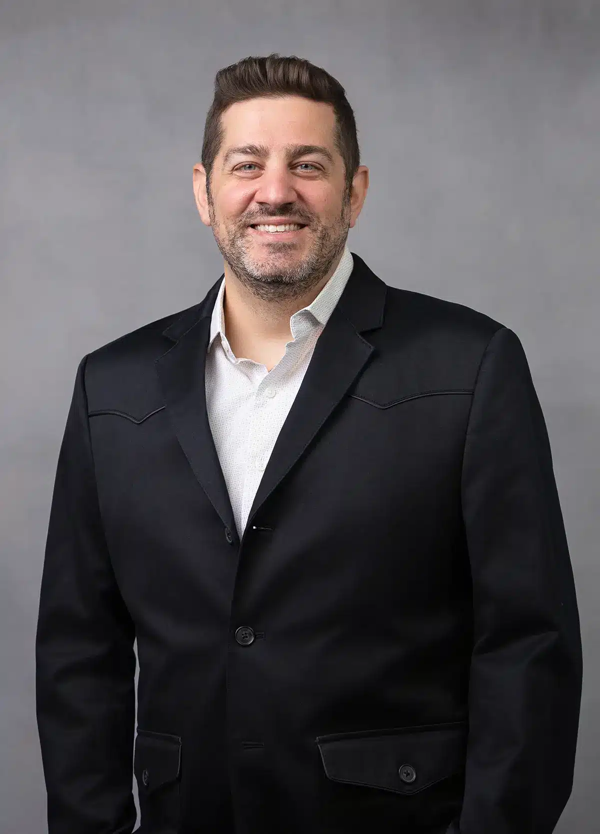 A man with short brown hair and a beard is smiling, wearing a black suit jacket over a white dress shirt, standing in front of a gray background.