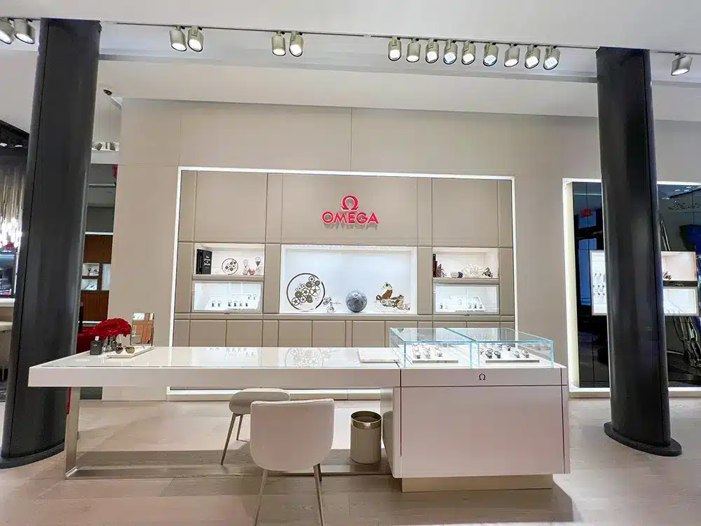 A modern, well-lit Omega watch boutique featuring display cases with luxury watches, a white counter with a chair, decorative red flowers, and the Omega logo on the back wall between two black columns.