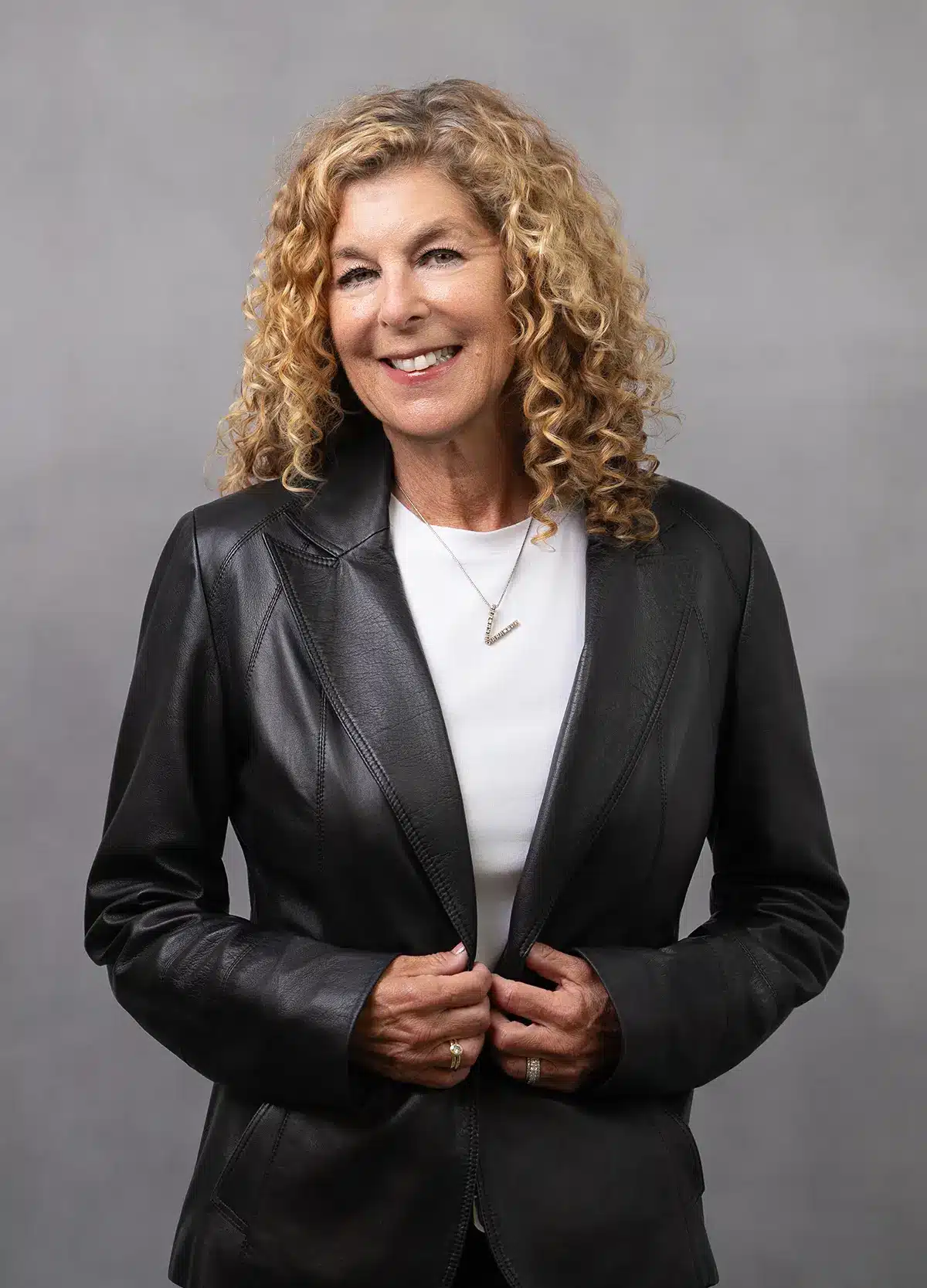 A woman with curly blonde hair smiles while wearing a black leather blazer over a white top, posing against a plain gray background with her hands holding the jacket.