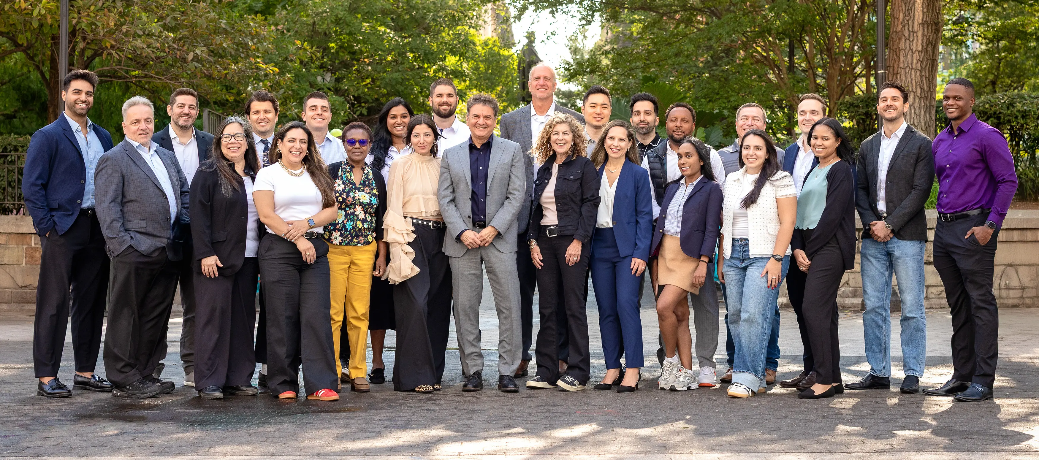A diverse group of 24 adults, dressed mostly in business or smart casual attire, stand outdoors in a park-like setting, smiling and posing together for a group photo. Trees and greenery are visible in the background.