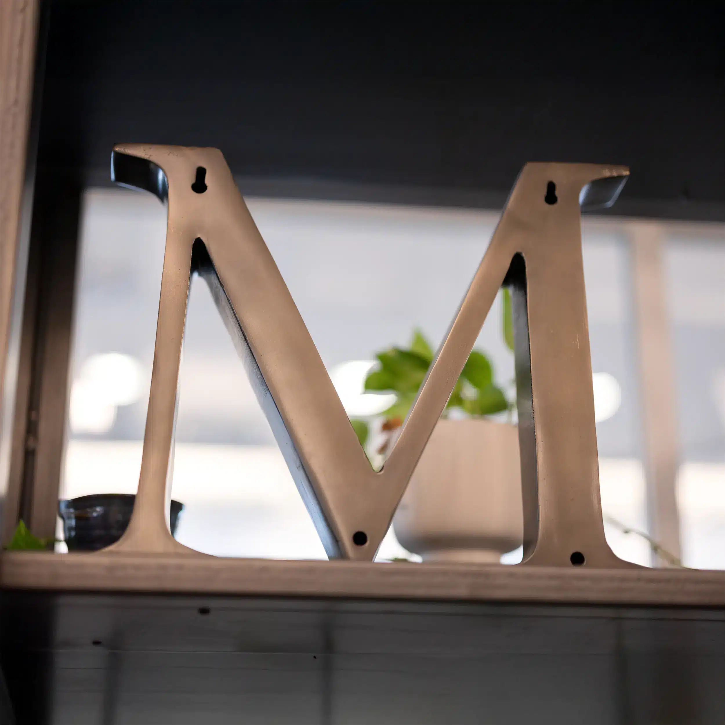 A metallic letter "M" sits on a shelf with a blurred green plant in a white pot in the background and soft natural light coming through a window.