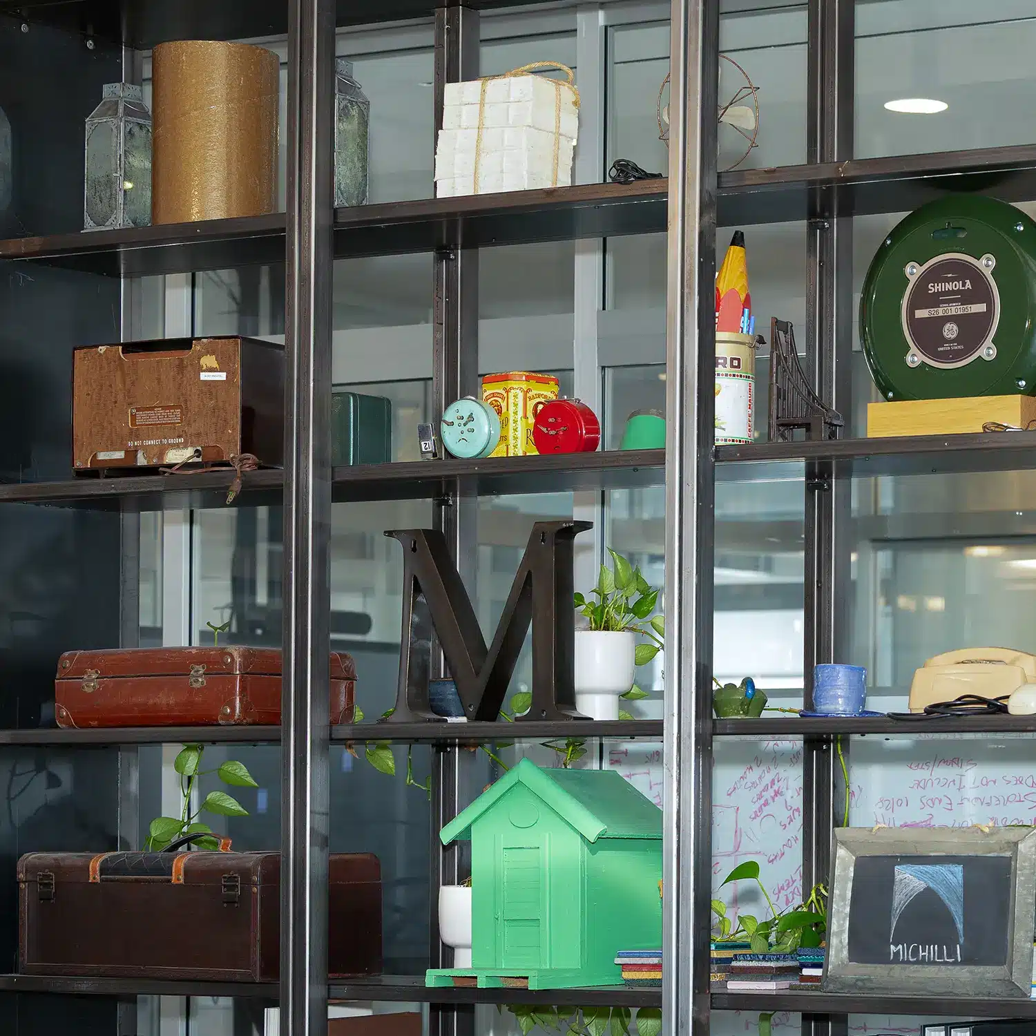 A black metal shelf filled with various items including vintage suitcases, potted plants, books, colorful decor pieces, a large letter M, a green model house, and assorted office supplies.