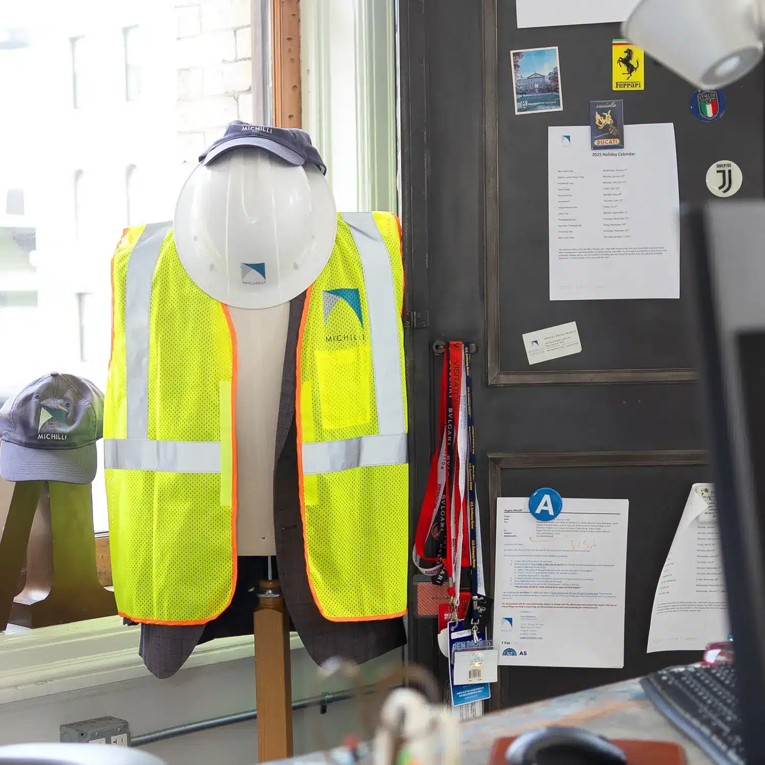 A mannequin wearing a bright yellow safety vest and white hard hat stands by a window in an office. Nearby, ID badges hang on a door covered with papers, notices, and photos.