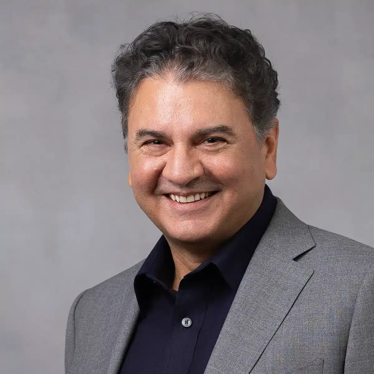 A man with short, curly dark hair smiles while wearing a gray suit jacket and a dark shirt, posing in front of a plain light gray background.