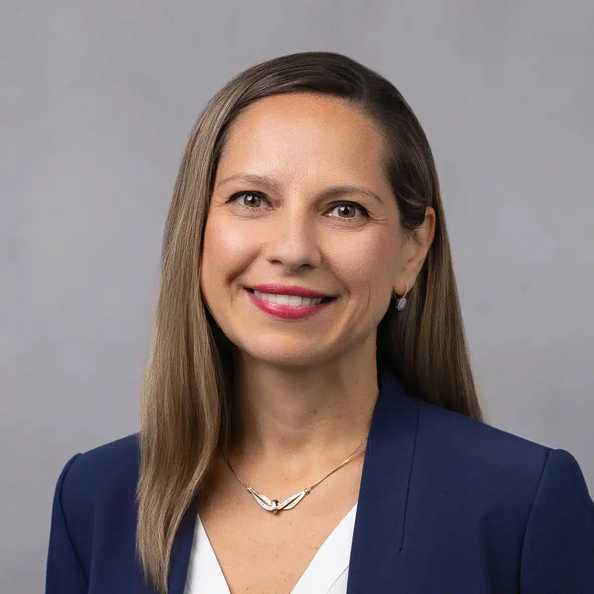 A woman with straight, shoulder-length light brown hair, wearing a navy blazer, white top, and a gold necklace, smiles at the camera against a plain gray background.