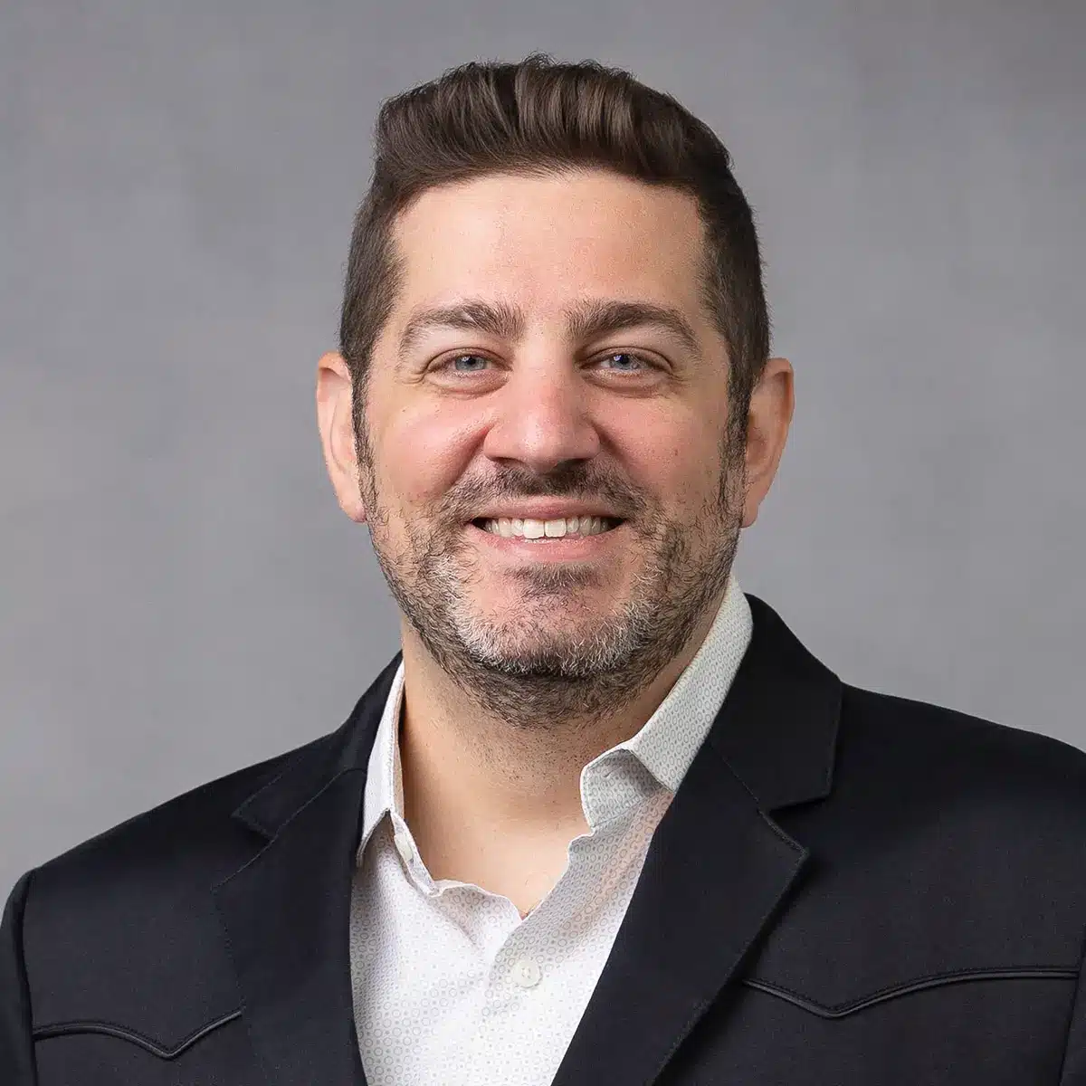 A man with short brown hair and a trimmed beard smiles at the camera. He is wearing a white shirt and a dark blazer, posing in front of a plain gray background.