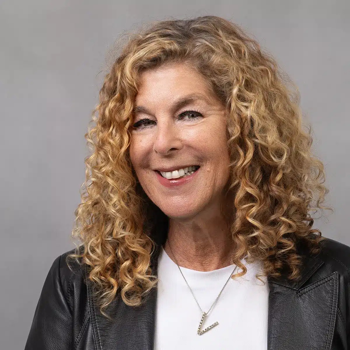 A woman with shoulder-length curly blonde hair smiles at the camera. She is wearing a black leather jacket over a white top and a necklace with a small pendant. The background is plain and light gray.