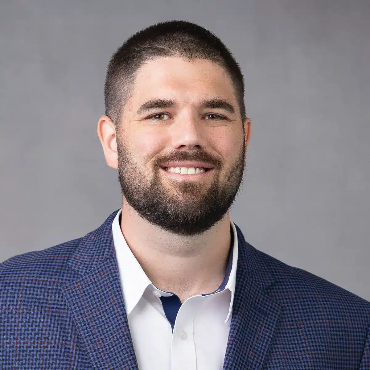 A man with short dark hair and a beard, smiling, wearing a white collared shirt and a blue checkered blazer, posed against a plain gray background.