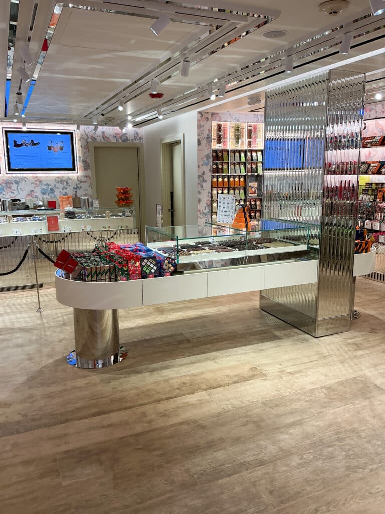 A brightly lit, modern candy store with a glass display case filled with assorted chocolates and treats, shelves of colorful packaged sweets, and a digital screen on the wall. The decor is sleek with floral accents.