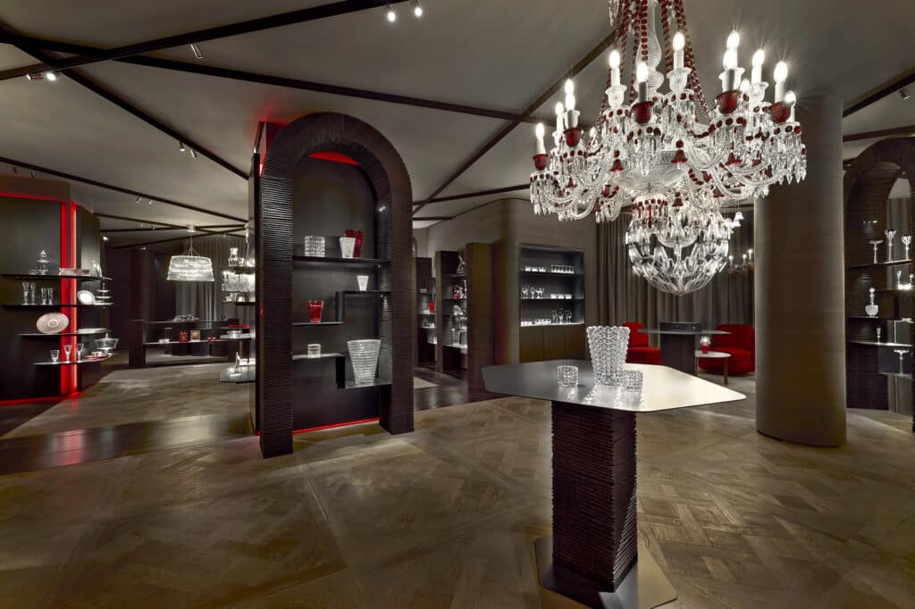 Luxurious showroom with glass display shelves holding crystal vases and glassware, ornate chandeliers with red accents, a central table with vases, and elegant lighting highlighting the room’s sophisticated decor.