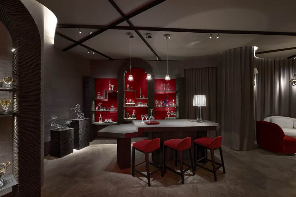A modern home bar with red shelves displaying glassware and bottles, three red barstools, a geometric bar counter, pendant lights, a silver sculpture, and taupe walls with dramatic lighting.
