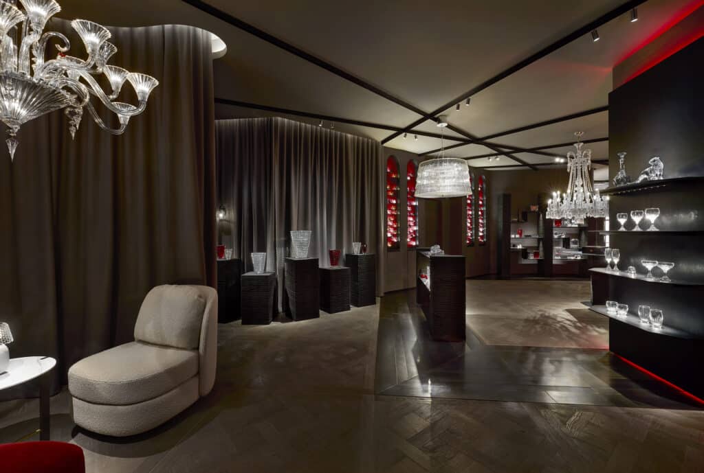 A luxurious interior showroom features elegant glassware displays, chandeliers, soft brown drapes, dark wood floors, and accent lighting, creating a sophisticated and inviting atmosphere.