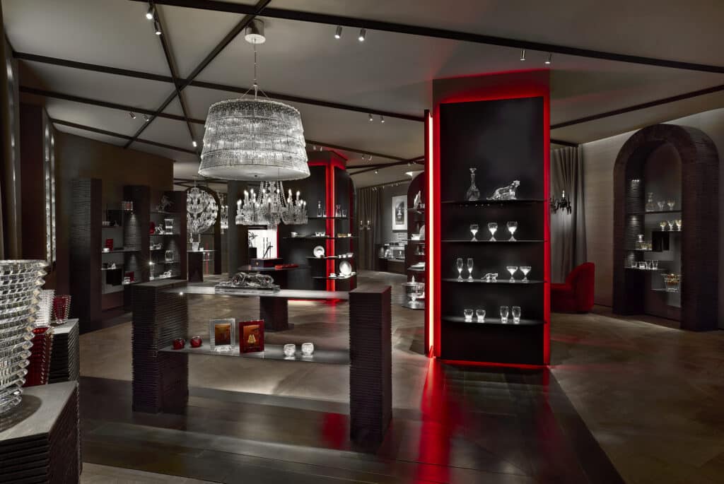 A modern, luxurious showroom displays glassware and crystal items on dark shelves, with red accent lighting and elegant chandeliers hanging from the ceiling, creating a sophisticated ambiance.