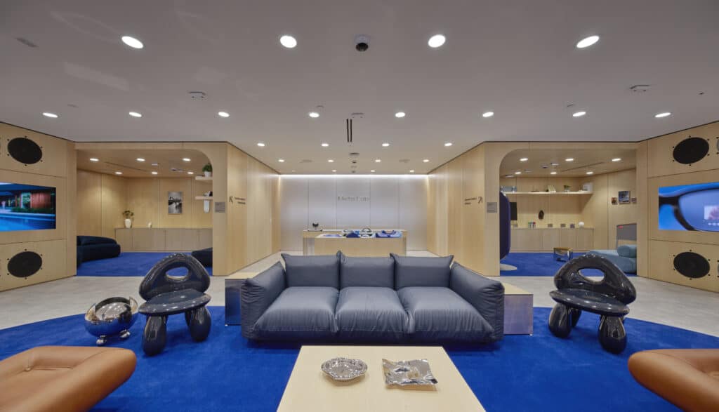 Modern lounge with blue carpet, grey sofa, two black sculptural chairs, and wooden walls featuring shelves and decor items. Recessed lighting brightens the space with a reception desk in the background.