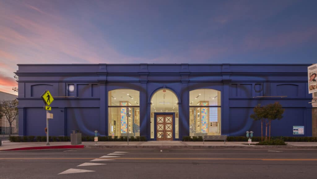 A modern blue building with large windows, a central arched entrance, and colorful displays inside, set against a twilight sky on a quiet city street.