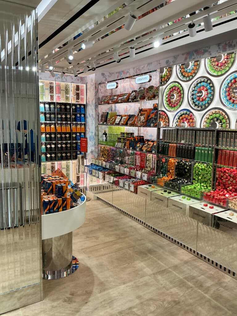 A brightly lit, modern candy store with colorful shelves filled with assorted sweets, chocolates, and gift boxes. The back wall features circular displays of macarons in various colors. Mirrored panels add a sleek touch.