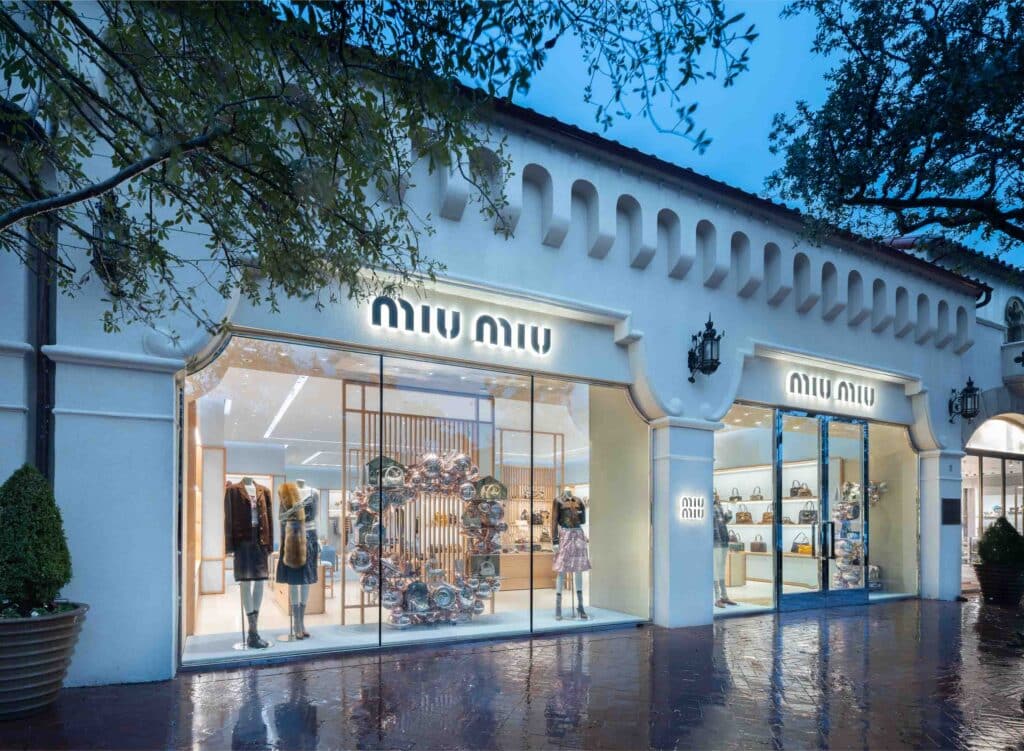Exterior view of a Miu Miu store at dusk, with large windows displaying clothing, mannequins, and bags inside. The building has white walls, tiled roof, and warm lights reflecting on wet pavement.