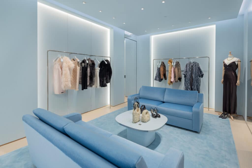 A modern boutique with light blue walls, two matching blue sofas, a blue rug, a white coffee table with bags, and clothing racks displaying dresses and outfits. A mannequin in a black dress stands in the corner.