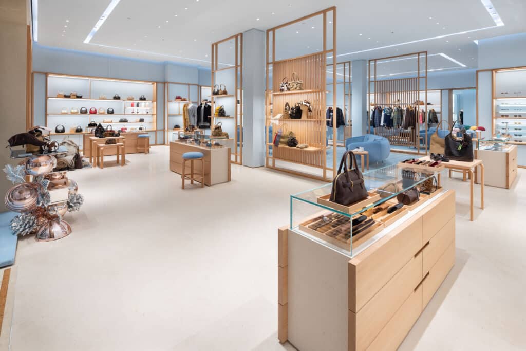 A modern, upscale boutique with light wood and glass displays featuring handbags, accessories, shoes, and clothing. The space is bright, open, and neatly organized, with soft lighting and minimalistic decor.