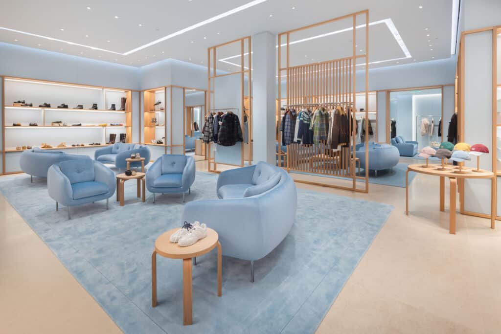 A modern boutique with soft blue armchairs, a matching rug, light wood accents, shelves displaying shoes, racks of clothes, and hats arranged on a round table. The space is bright, open, and neatly organized.