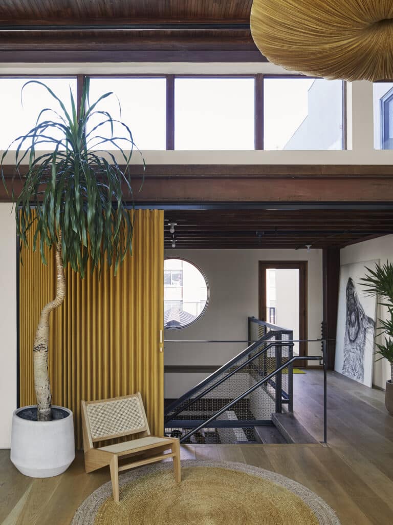 A modern interior with wooden floors, a tall potted plant, a woven chair, a round rug, a yellow corrugated divider, large windows, and a metal staircase. A round window and artwork are visible in the background.
