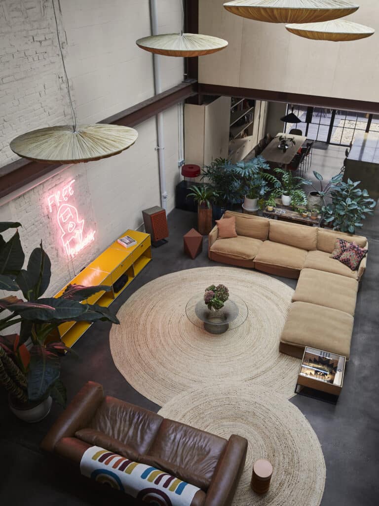A modern loft living room with a large tan sectional sofa, a brown leather couch, a round beige rug, plants, a neon 