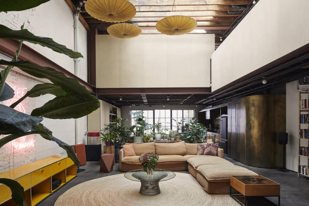 A modern loft living room with exposed beams, large tan sectional sofa, round woven rug, indoor plants, a yellow sideboard, and warm lighting. Large windows at the back let in natural light.